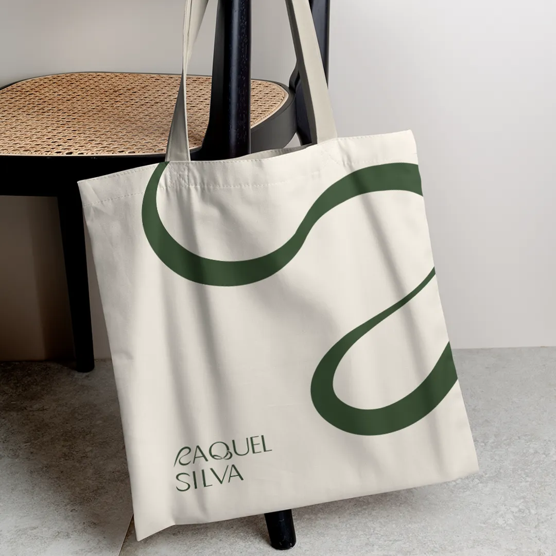 Mockup de uma ecobag bege pendurada numa cadeira, com o logotipo 'Raquel Silva' em verde, criado por Manuel Silva Design. A composição moderna e elegante valoriza a marca e transmite sofisticação e identidade visual sustentável.